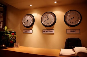 Tres relojes en una pared con la etiqueta 'NEW-YORK', 'Le Ruisseau' y 'SYDNEY', con un escritorio y una silla al frente.