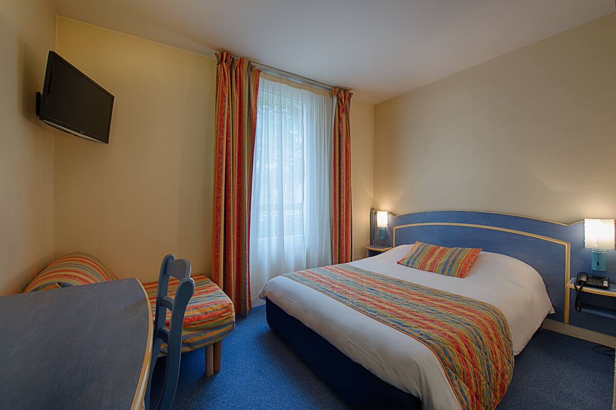 A hotel room with a bed, a desk, a chair, a TV mounted on the wall, and a window with curtains.
