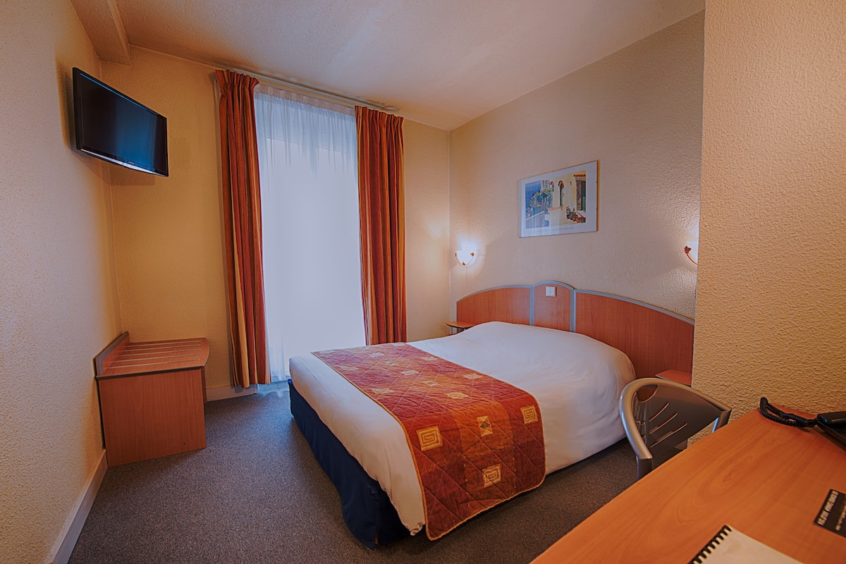 A hotel room with a bed, a desk, a chair, a television, and a painting on the wall.