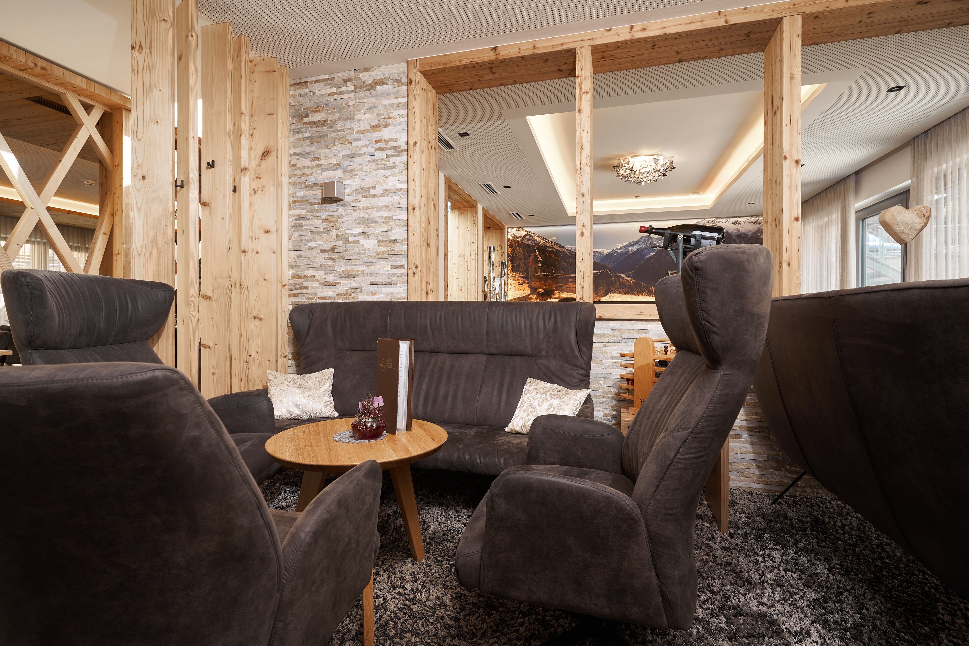 A cozy lounge area with dark sofas, a round wooden table, and a mountain view through a large window.