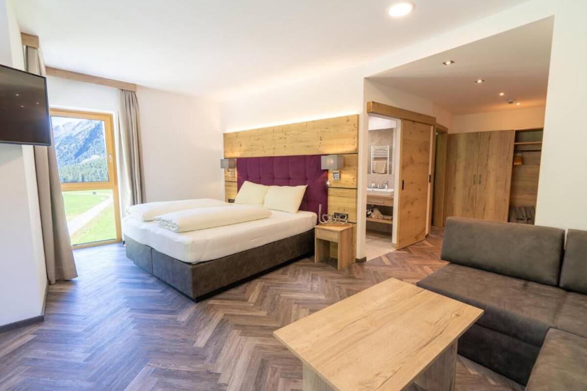 A modern hotel room with a large bed, a wooden headboard, a sofa, a coffee table, and a TV.