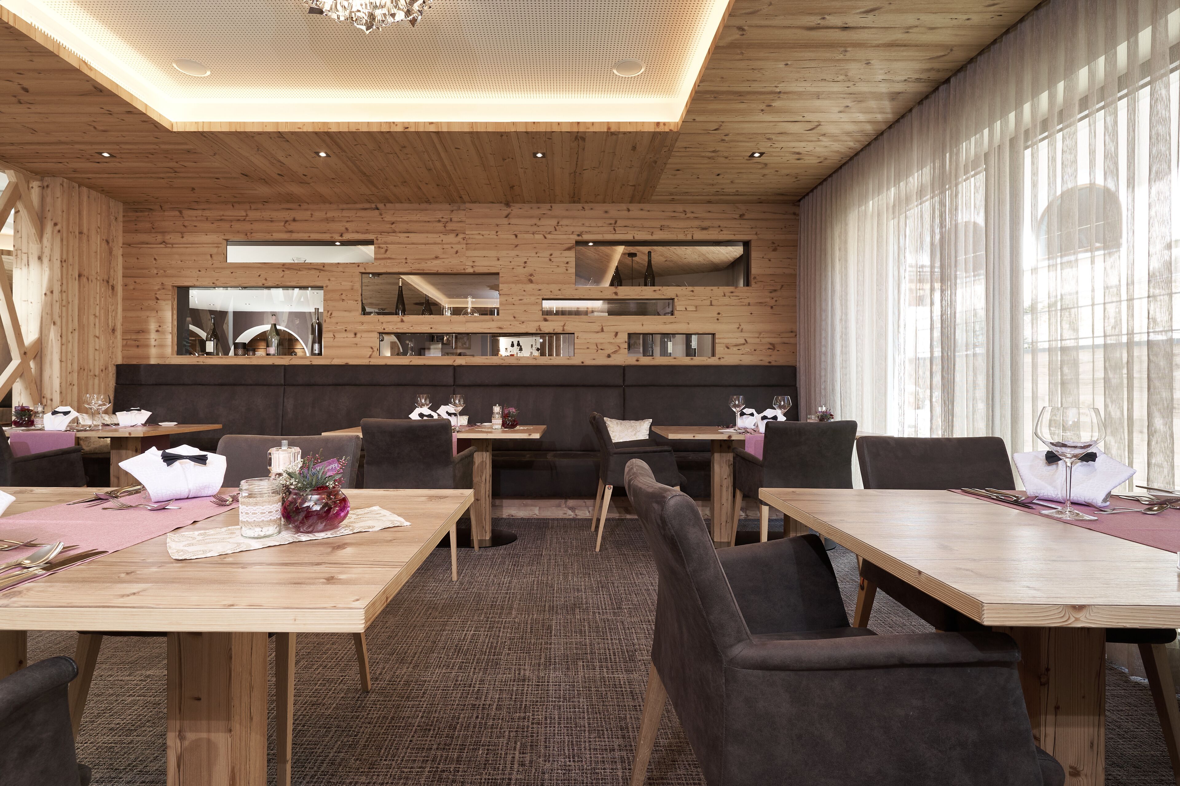 A modern dining area with wooden tables and chairs, a central bar area, and large windows allowing natural light.