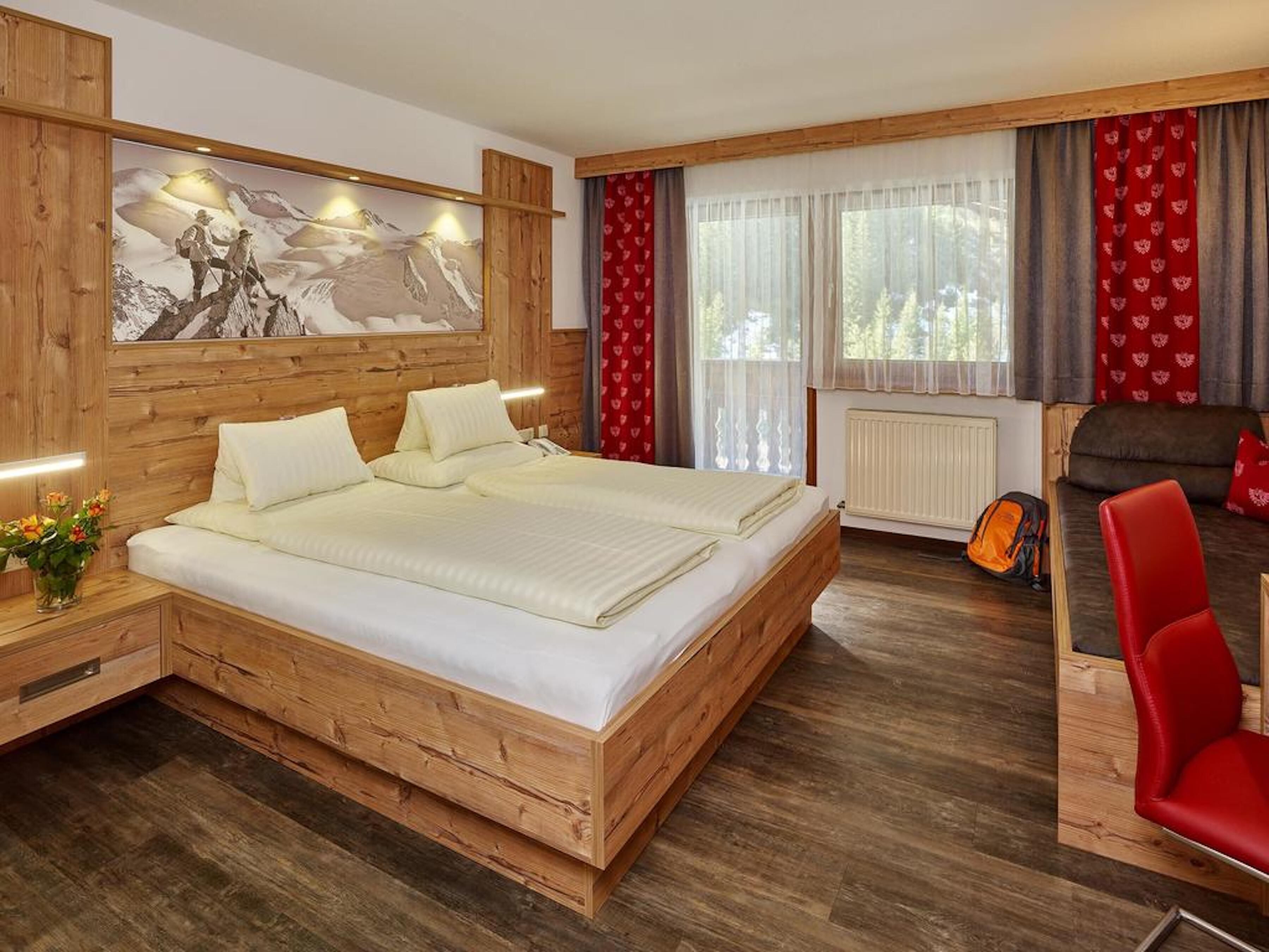 A wooden-framed double bed with white linens, a wooden nightstand with a vase of flowers, a red chair, and a large mountain-themed painting on the wall.