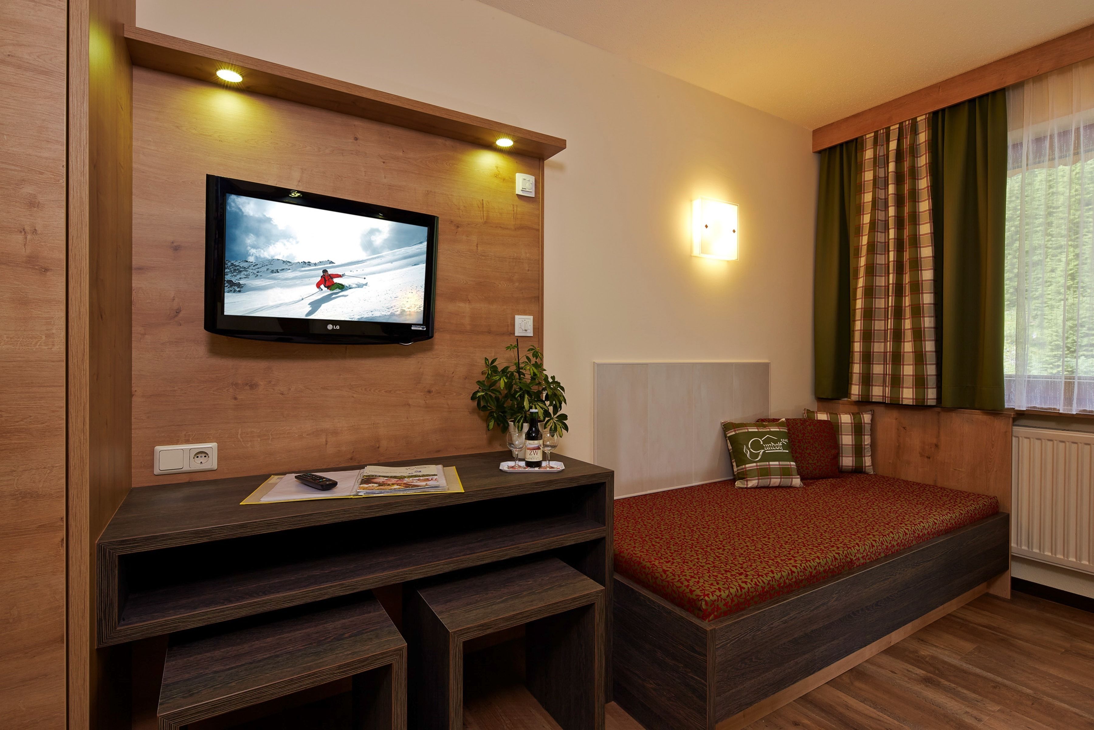 Ein Zimmer mit an der Wand montiertem Holz-Fernseher, einem kleinen Tisch mit Pflanze und Fernbedienung, einem Bett mit rot gemustertem Überwurf und einem Fenster mit grünen Vorhängen.