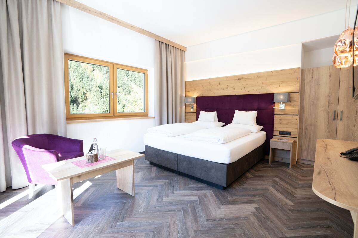 A modern hotel room with a large bed, a wooden headboard, a small table, a purple chair, and a view of greenery through the window.