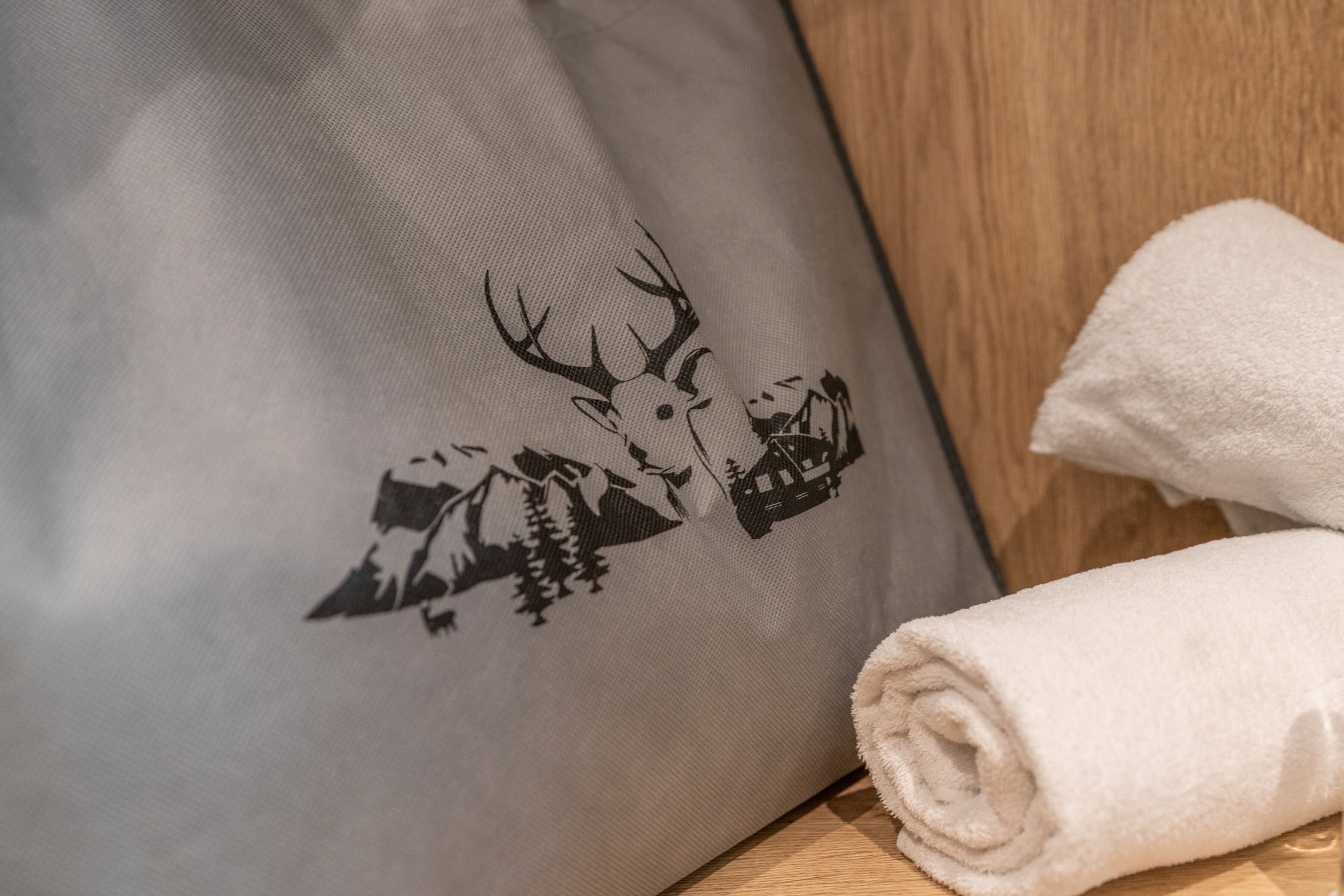 Un coussin orné d’un motif représentant un cerf et un chalet de montagne, une serviette blanche roulée et une surface en bois.