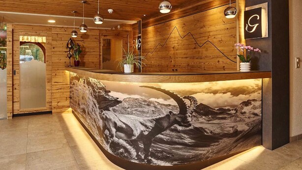 A reception desk with a wooden finish, a mountain landscape mural, and pendant lighting. - Hotel Gundolf  (Sankt Leonhard im Pitztal)