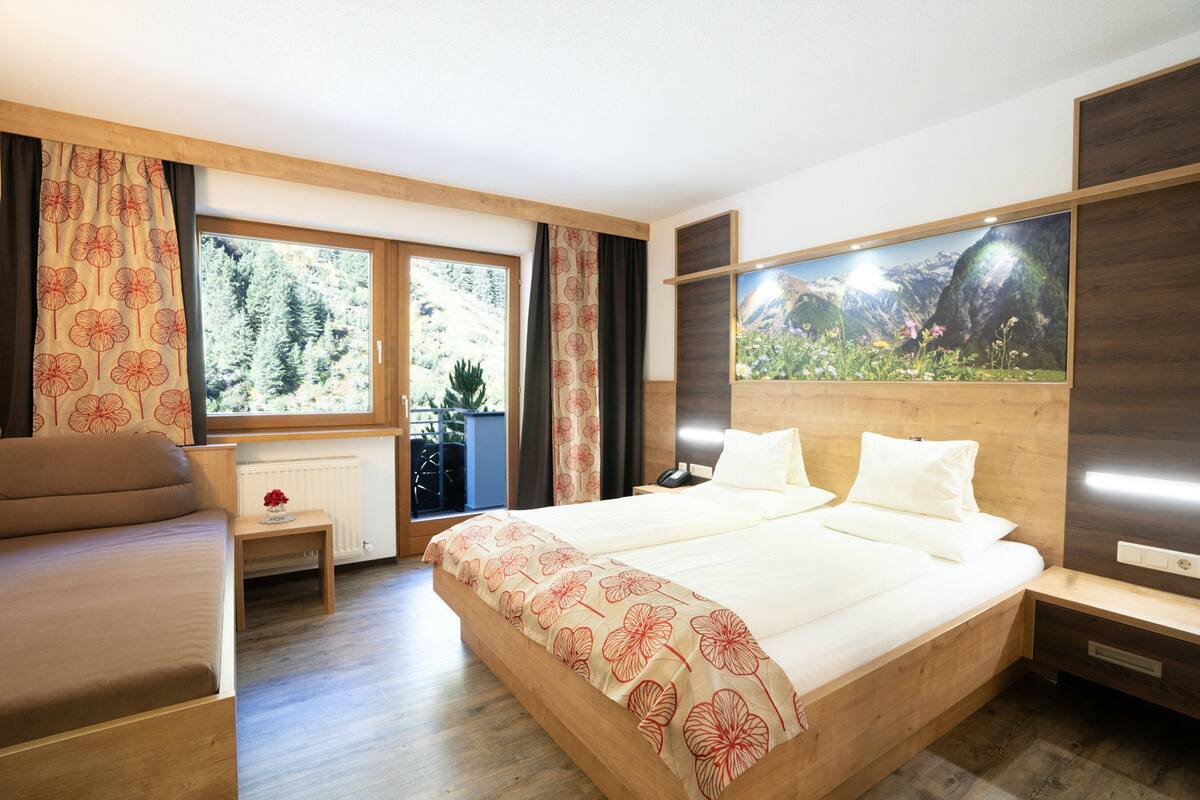 A bedroom with a large bed, a wooden headboard, a nightstand, a bedside table, a window with curtains, and a wall-mounted TV.