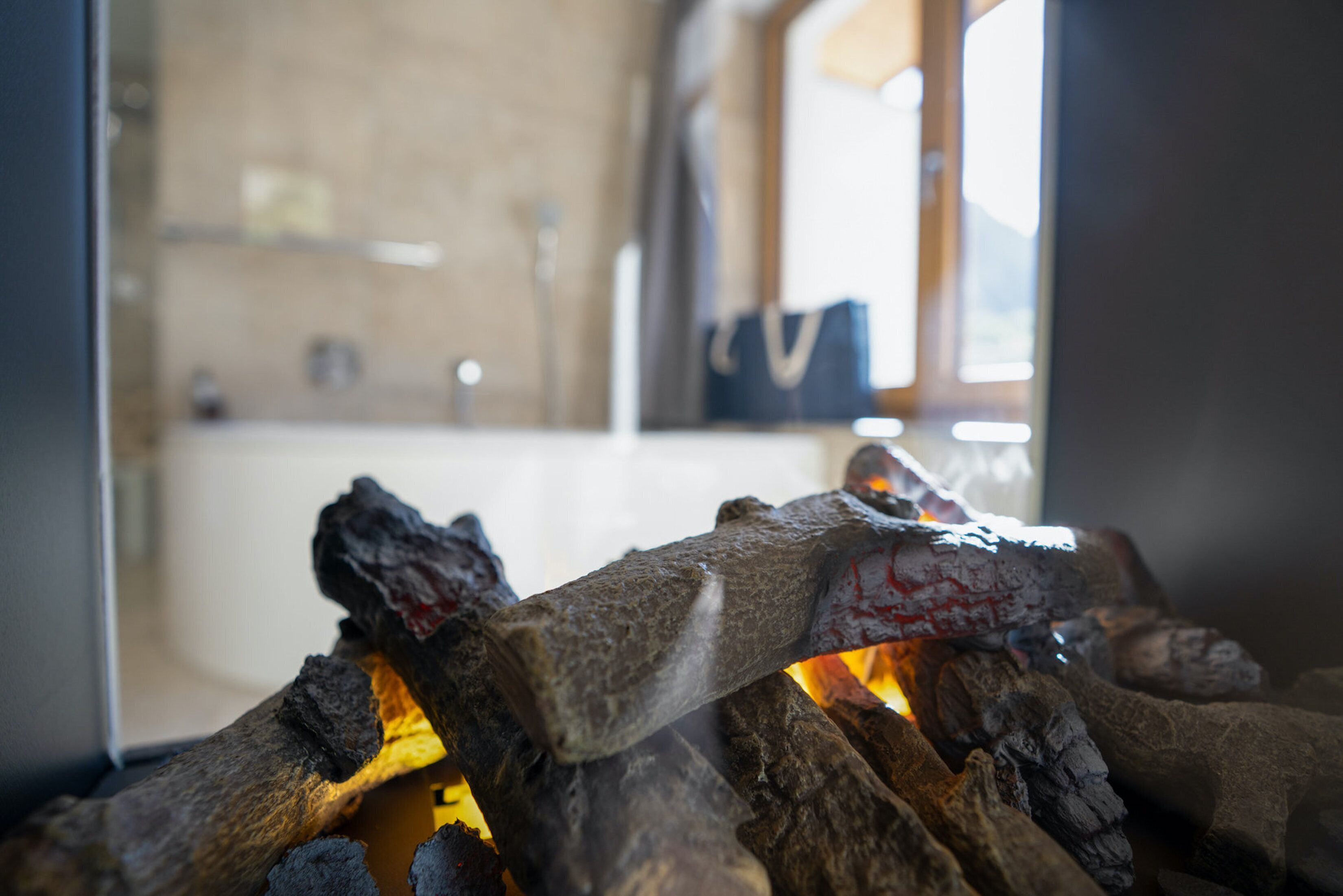 A close-up of burning wood in a fireplace, with a blurred background suggesting a room with a window and a wall.
