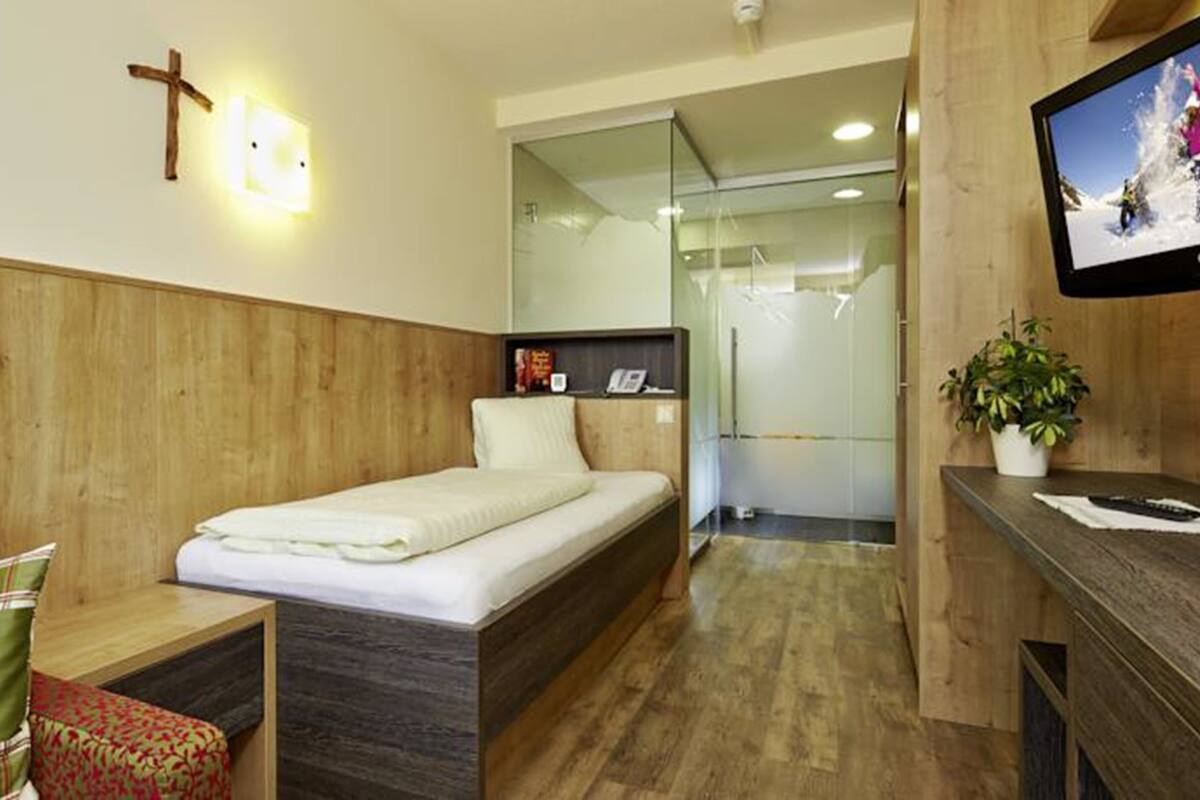 A hotel room with a bed, a small seating area with a chair, a television, and a cross on the wall.