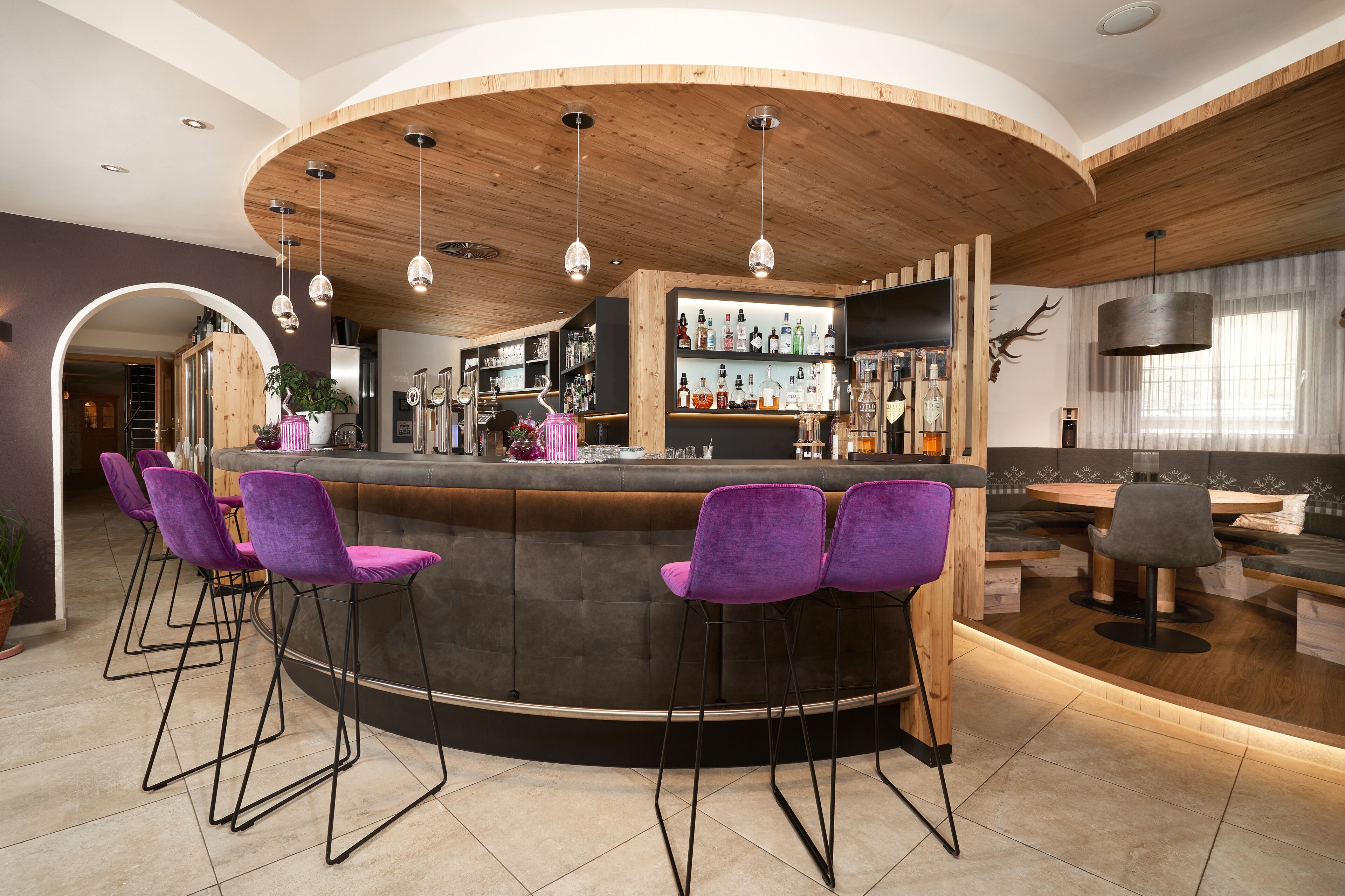 A bar area with a curved wooden counter, purple bar stools, and a variety of bottles on shelves.