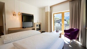 A hotel room with a large bed, a TV mounted on the wall, a chair, and a view of mountains through the window. - Hotel Gundolf  (Sankt Leonhard im Pitztal)