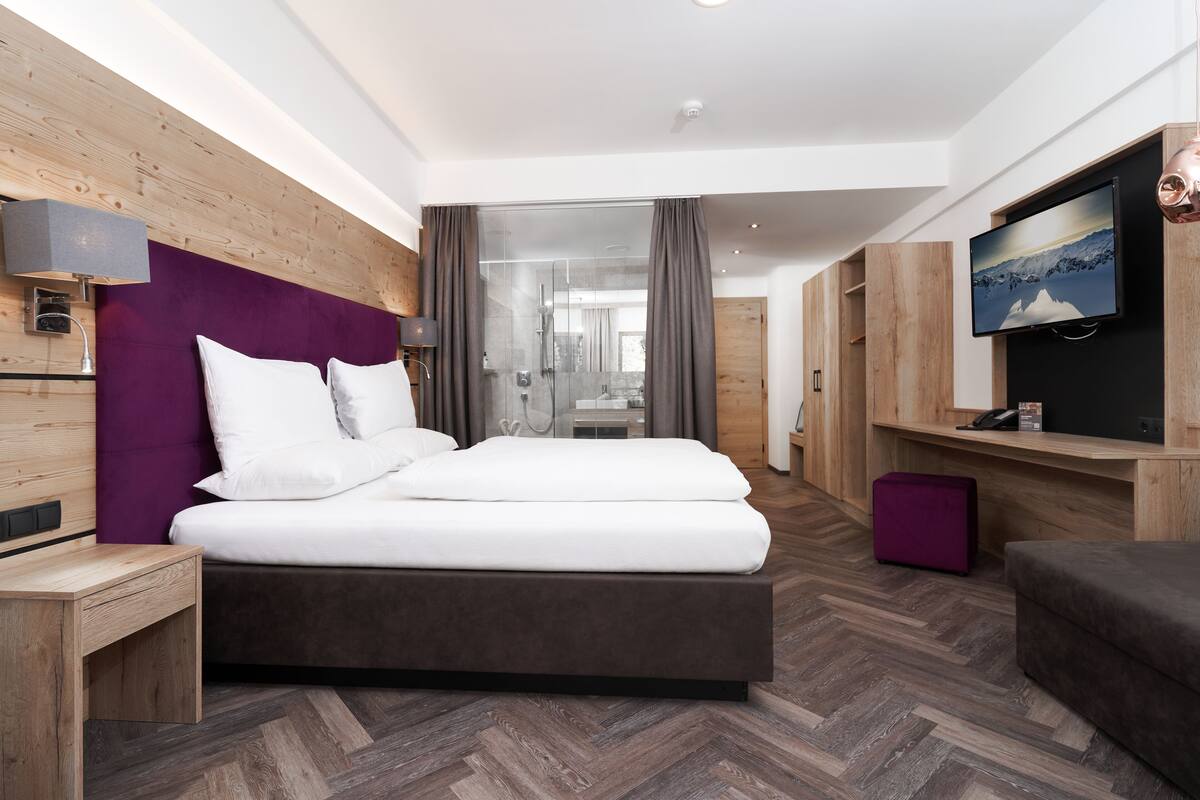 A modern hotel room with a large bed, wooden headboard, a TV, and a glass shower area.