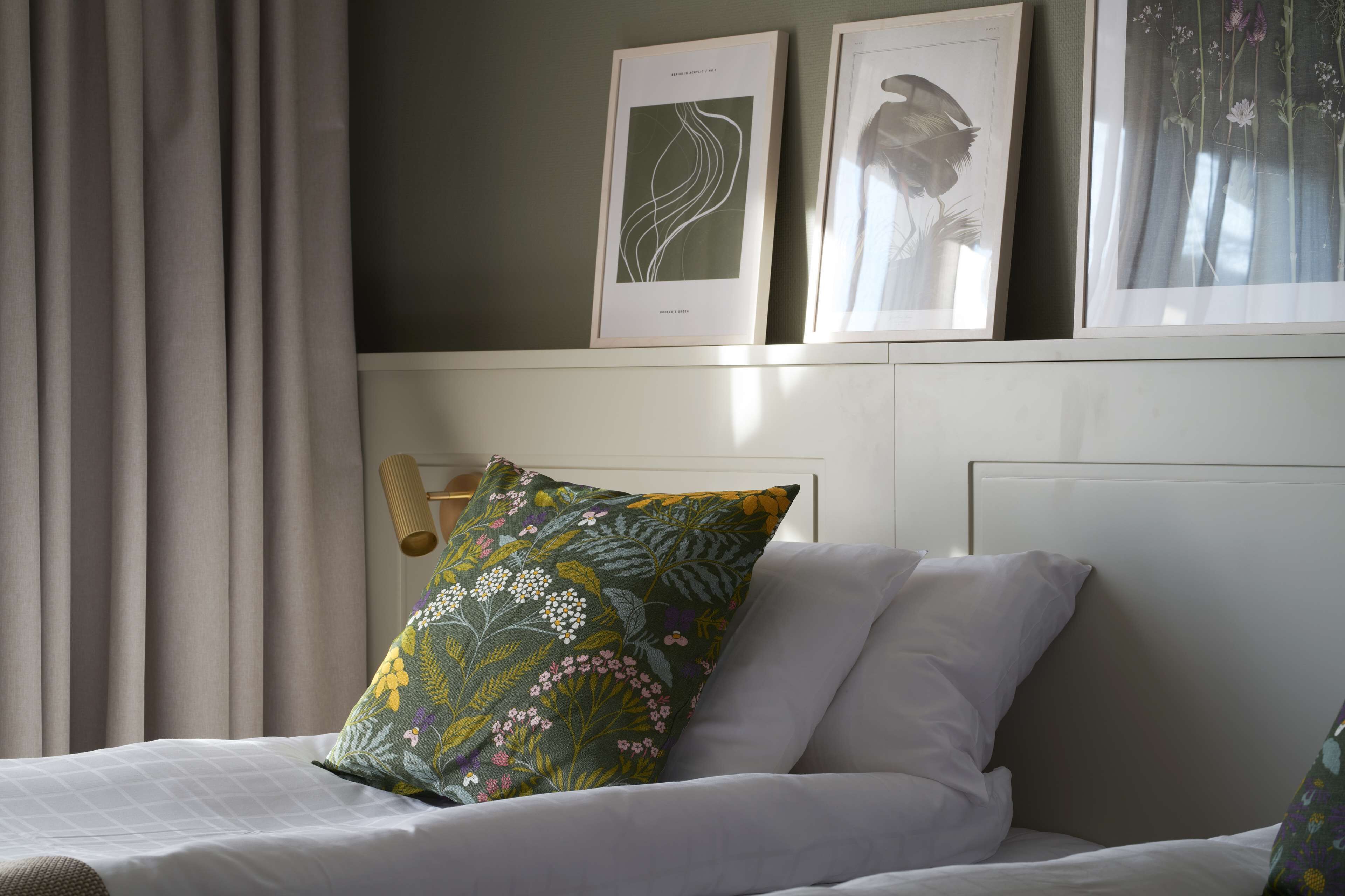 Una cama bien hecha con ropa de cama blanca y una almohada estampada con motivos florales, un grabado botánico enmarco y una cortina en una ventana.
