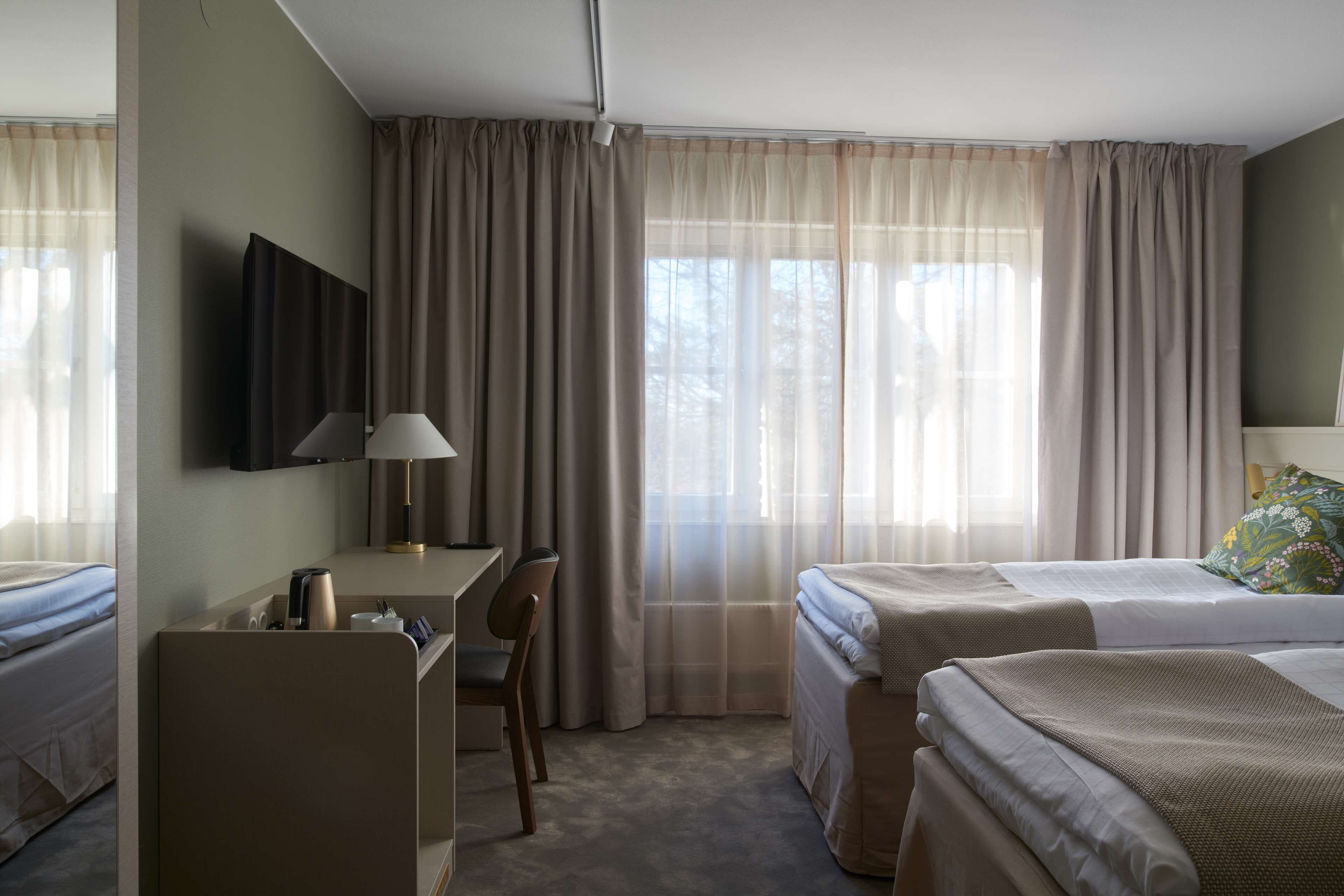 A hotel room with two beds, a desk, a chair, a TV, and large windows with curtains.