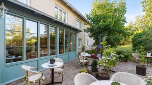 Een overdekt terras met rieten stoelen en tafels, omringd door potplanten en uitzicht op de tuin.