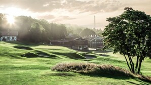 Een golfbaan met glooiende groene heuvels, een clubhuis en een hoge toren op de achtergrond.
