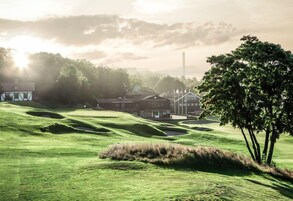 Un campo de golf con colinas verdes ondulantes, un club de campo y una torre alta al fondo.
