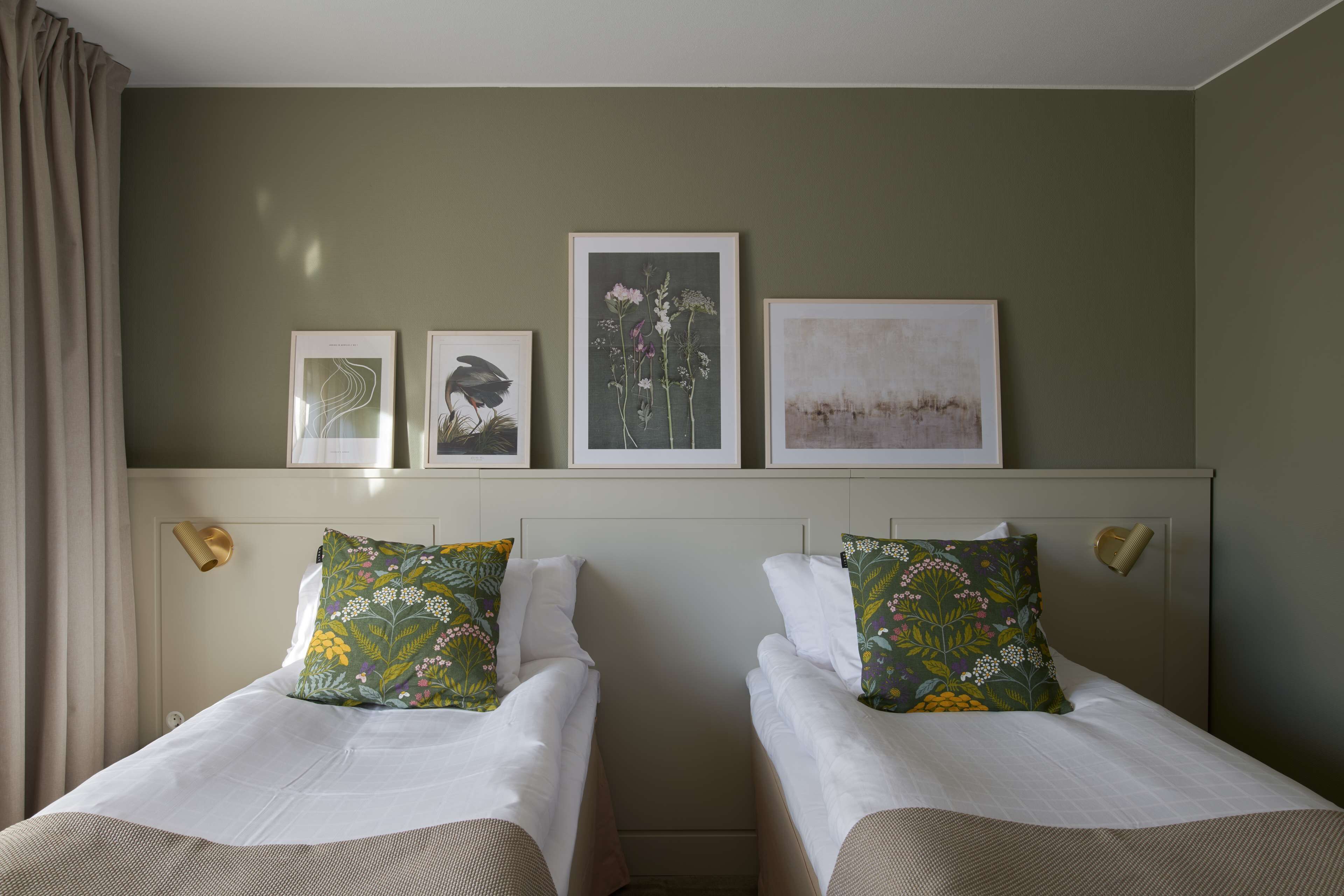 Ein Hotelzimmer mit zwei Betten, jedes mit blumigem Kissen, einem Kopfteil mit Wandleuchten und drei gerahmten Kunstwerken darüber.