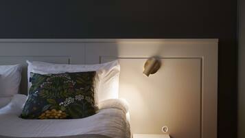 Una cama bien hecha con sábanas blancas y un cojín estampado con motivos florales, una mesita de noche con lámpara, y una lámpara de pared.