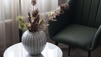 Un vase contenant des fleurs séchées est posé sur une table ronde, à côté d’un fauteuil recouvert d’un tissu vert.