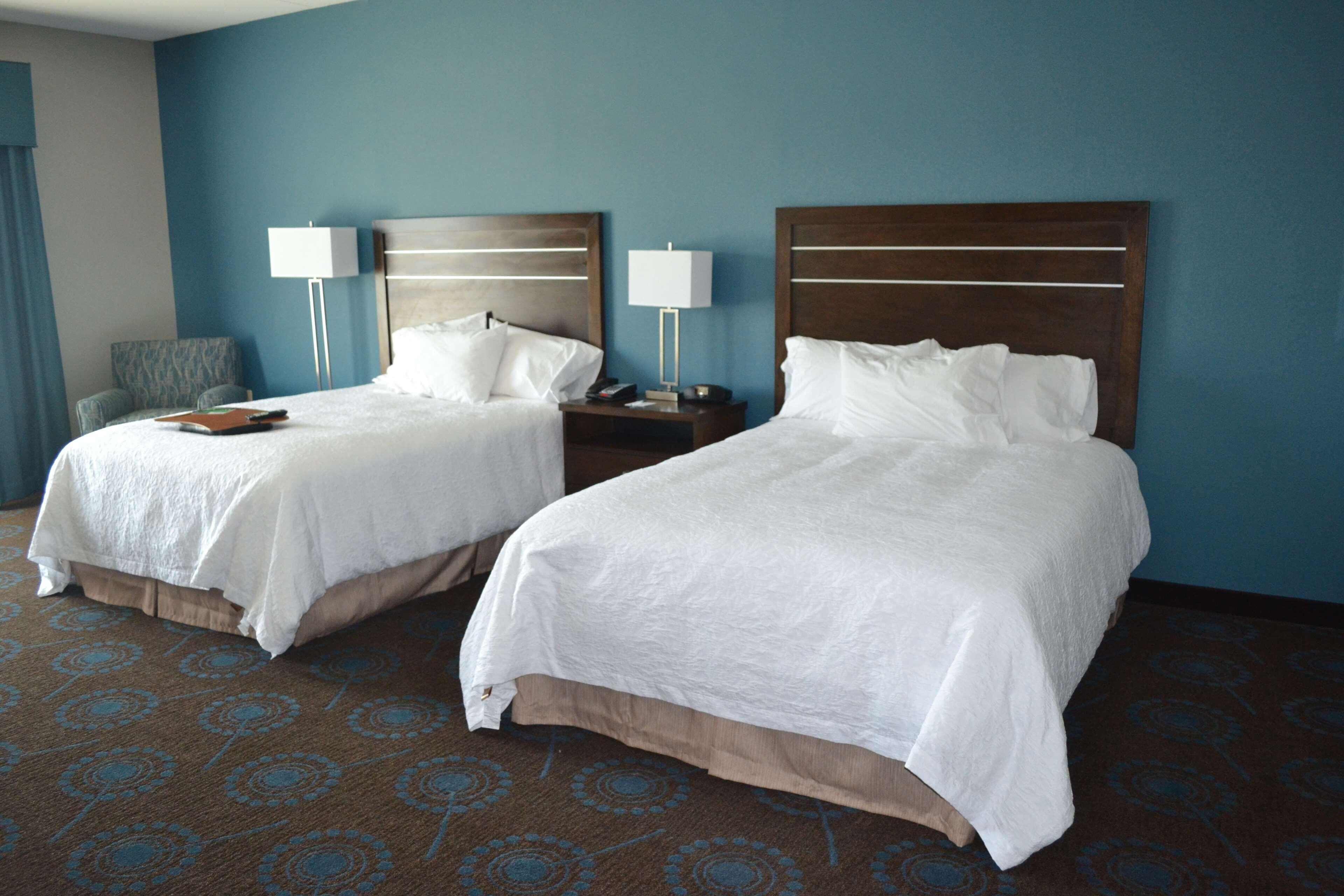 Two beds with white linens, a nightstand with a lamp, a chair, and a patterned carpet in a hotel room.