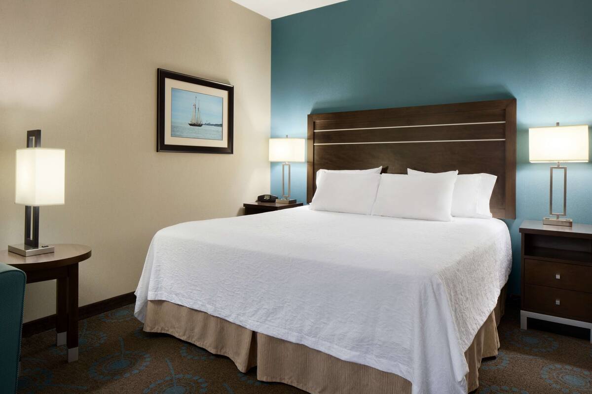 A hotel room with a large bed, two bedside tables with lamps, a framed picture on the wall, and a carpeted floor.
