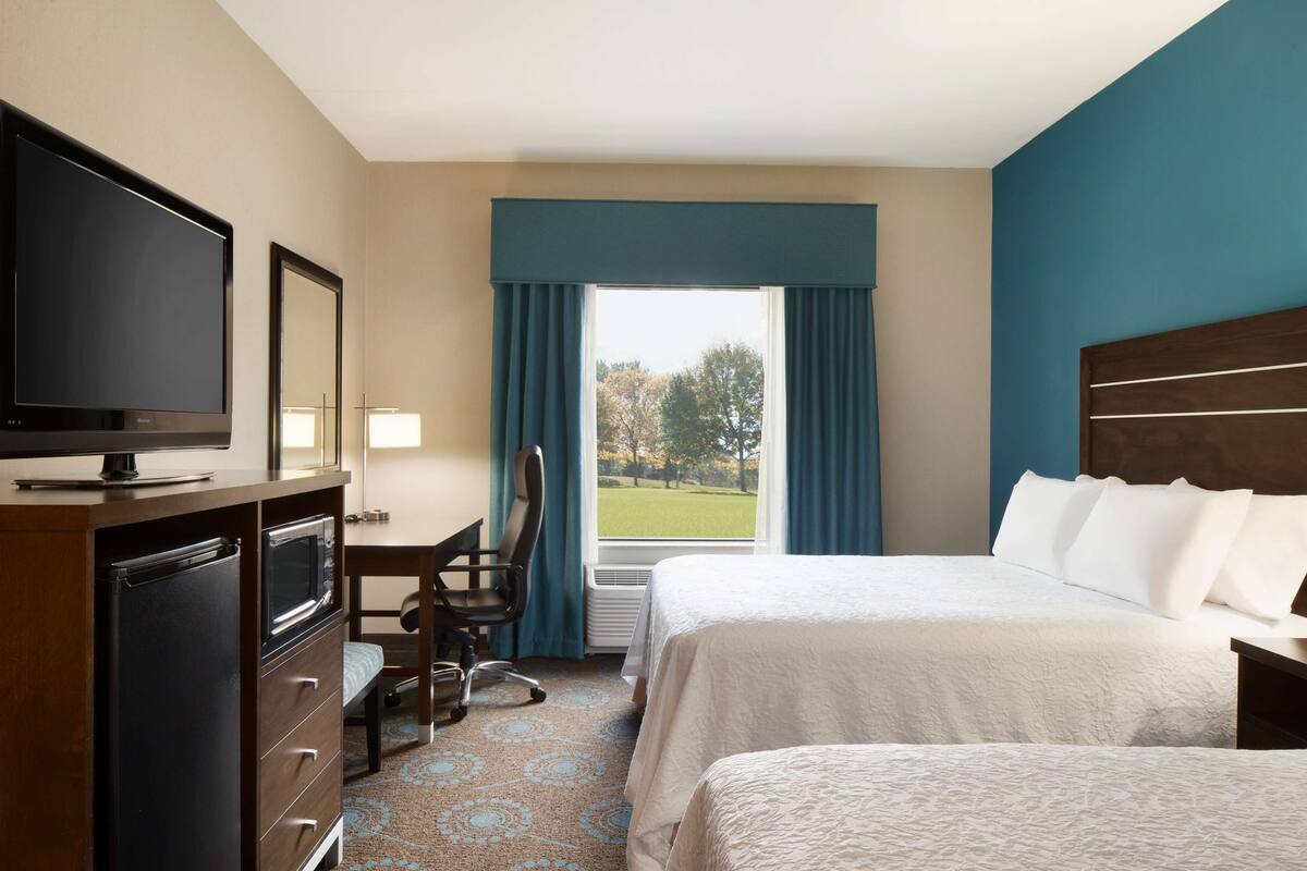 A hotel room with a bed, a desk with a chair, a television, and a window with curtains.