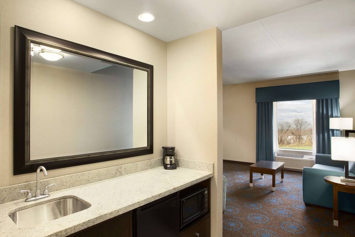 A hotel room with a large mirror, a sink, a coffee maker, a microwave, a small table, a couch, and a window with curtains.