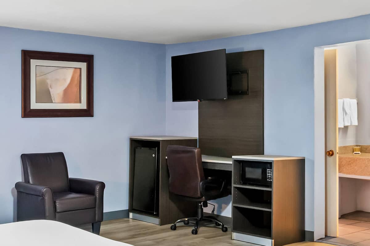 A hotel room with a bed, a desk with a computer, a chair, a TV mounted on the wall, and a bathroom visible through an open door.