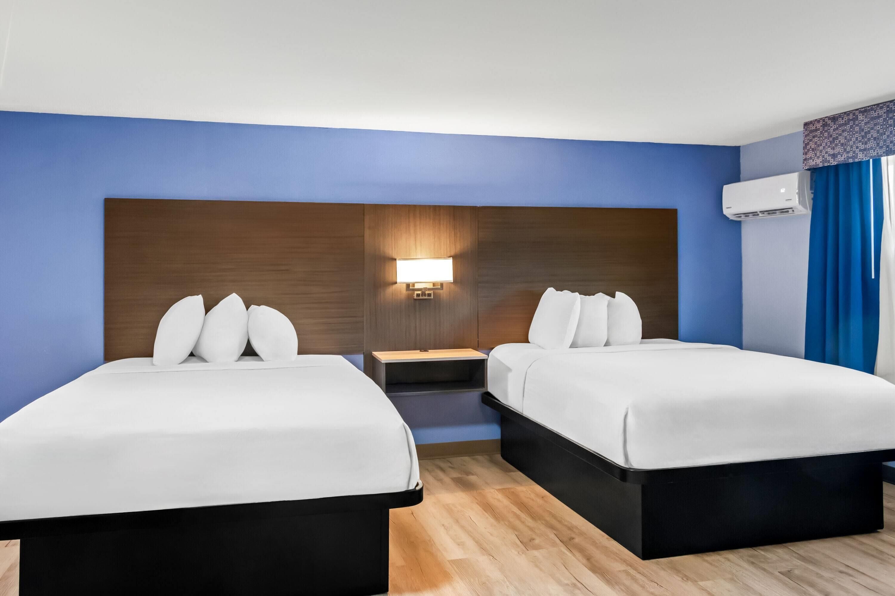 A hotel room with two beds, a nightstand, a wall-mounted lamp, and an air conditioning unit.