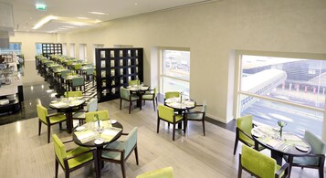 Un área de comedor moderna con varias mesas y sillas, un separador central negro y ventanas grandes que ofrecen vistas del paisaje urbano.