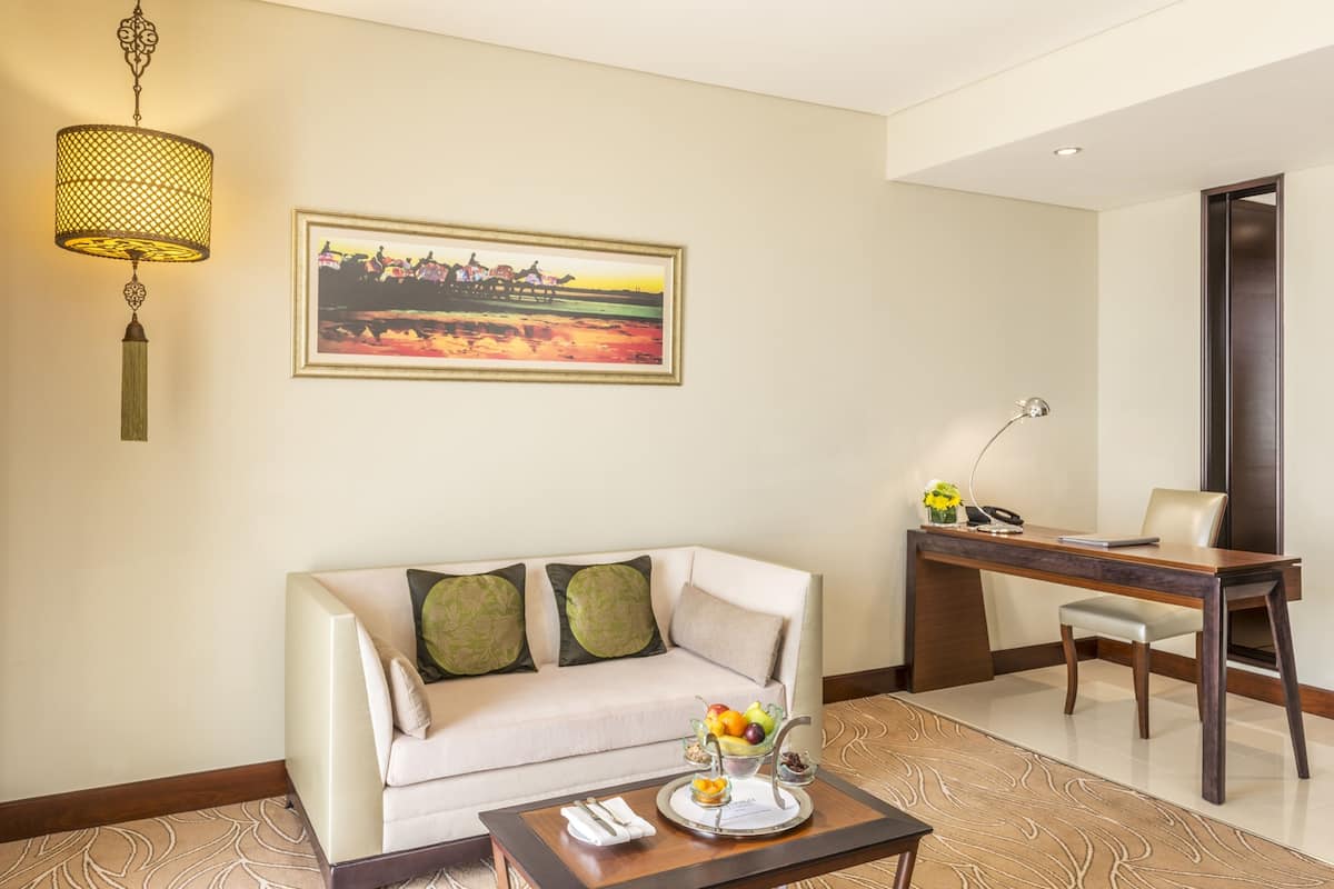 Une chambre d’hôtel équipée d’un canapé, d’une table à manger, d’un bureau, d’une lampe, d’un tableau et d’un panier de fruits.