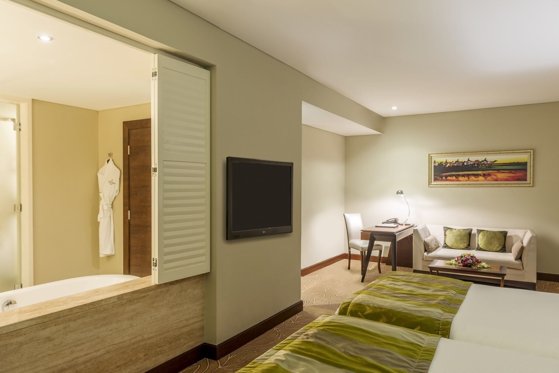 A hotel room with a large mirror, a flat-screen TV, a desk with a chair, a sofa, and a bed with green bedding.