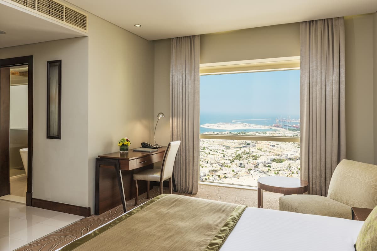 Une chambre d’hôtel équipée d’un lit, d’un bureau, d’une chaise, d’une petite table et offrant une vue sur le paysage urbain et la côte.