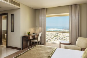 A hotel room with a bed, a desk, a chair, a small table, and a view of a cityscape through large windows.