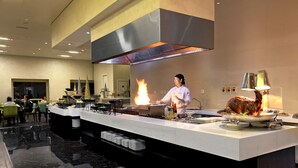 Une cuisine moderne où un chef prépare des plats, avec un grand îlot central et un buffet proposant divers mets.