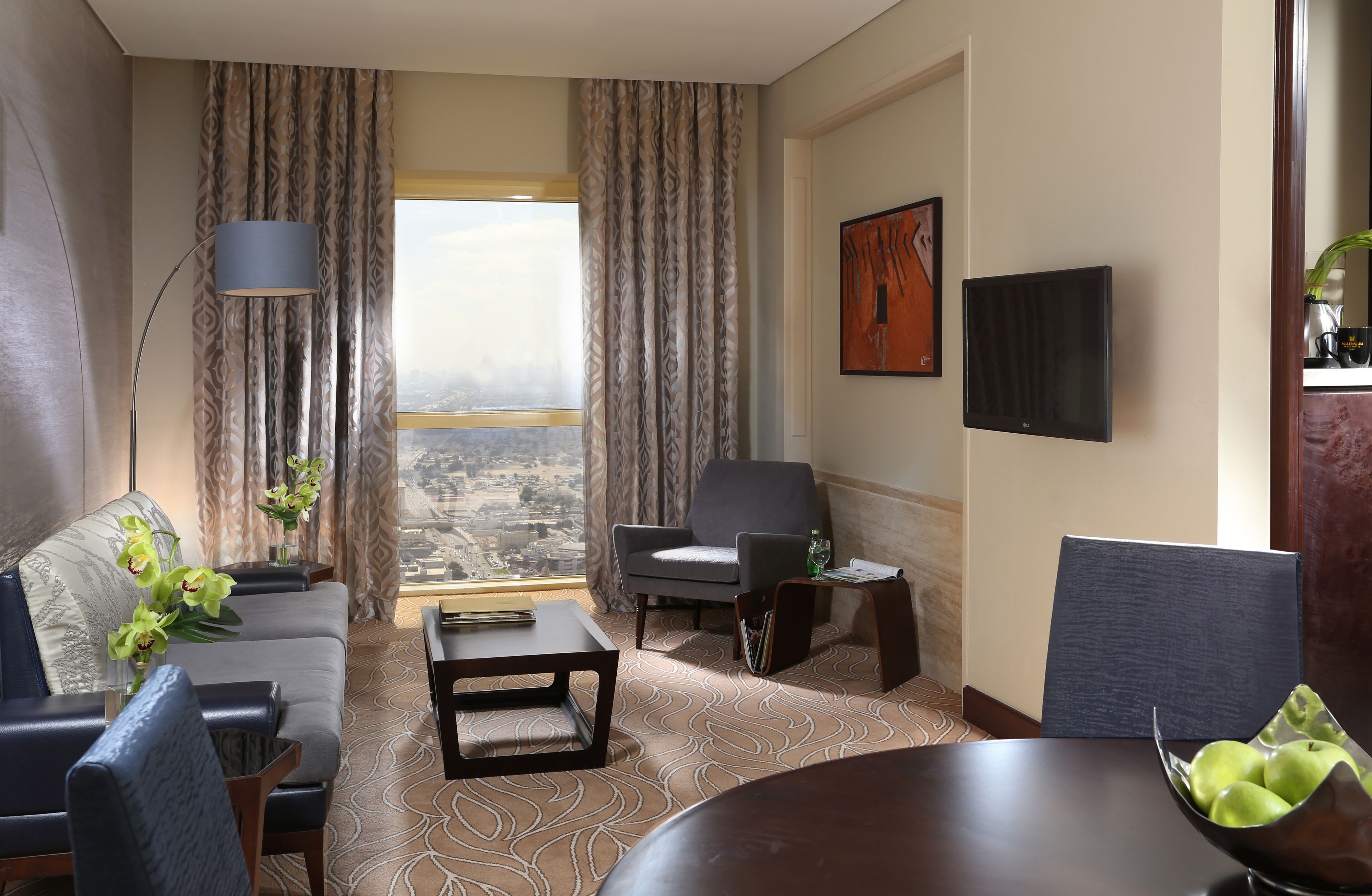 Habitación de hotel con sofá, sillones, una mesa de centro, una mesa de comedor con manzanas, un televisor y vistas a la ciudad a través de amplias ventanas.