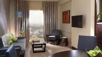 Habitación de hotel con sofá, sillones, una mesa de centro, una mesa de comedor con manzanas, un televisor y vistas a la ciudad a través de amplias ventanas.