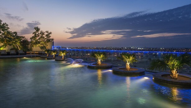 Vue nocturne d’une piscine sur le toit, agrémentée de jeux d’eau et de palmiers, d’où l’on aperçoit un paysage urbain et un pont.