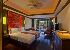 Amplia habitación de hotel con una cama grande, una zona de estar con mesa, televisión y un balcón con vistas a palmeras.