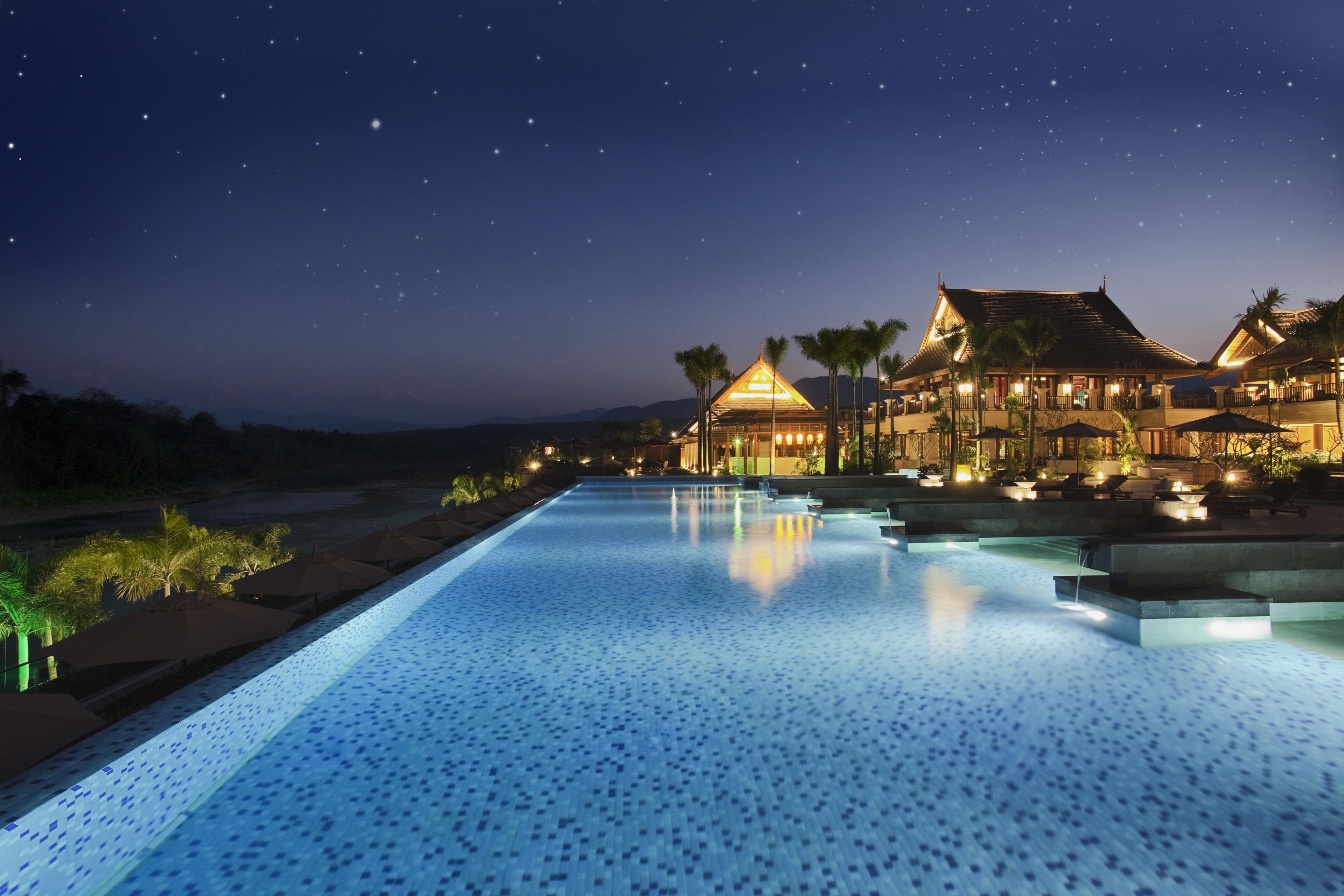 A nighttime view of a resort with a swimming pool, a building with a traditional roof, and palm trees.
