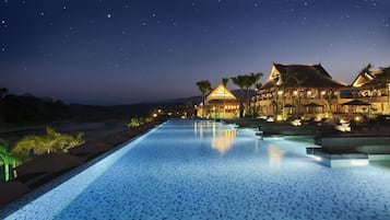 A nighttime view of a resort with a swimming pool, a building with a traditional roof, and palm trees.