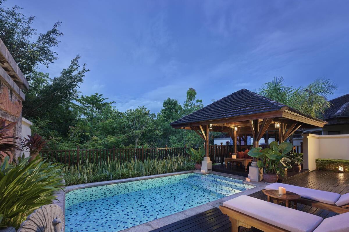A swimming pool with a tiled deck, lounge chairs, a gazebo, and lush greenery.