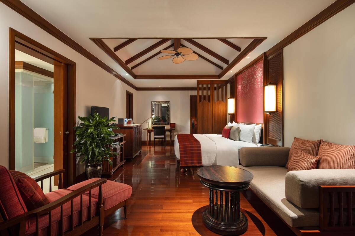 A hotel room with a wooden ceiling, a bed with a red and white blanket, a sofa, a chair, a table, and a potted plant.