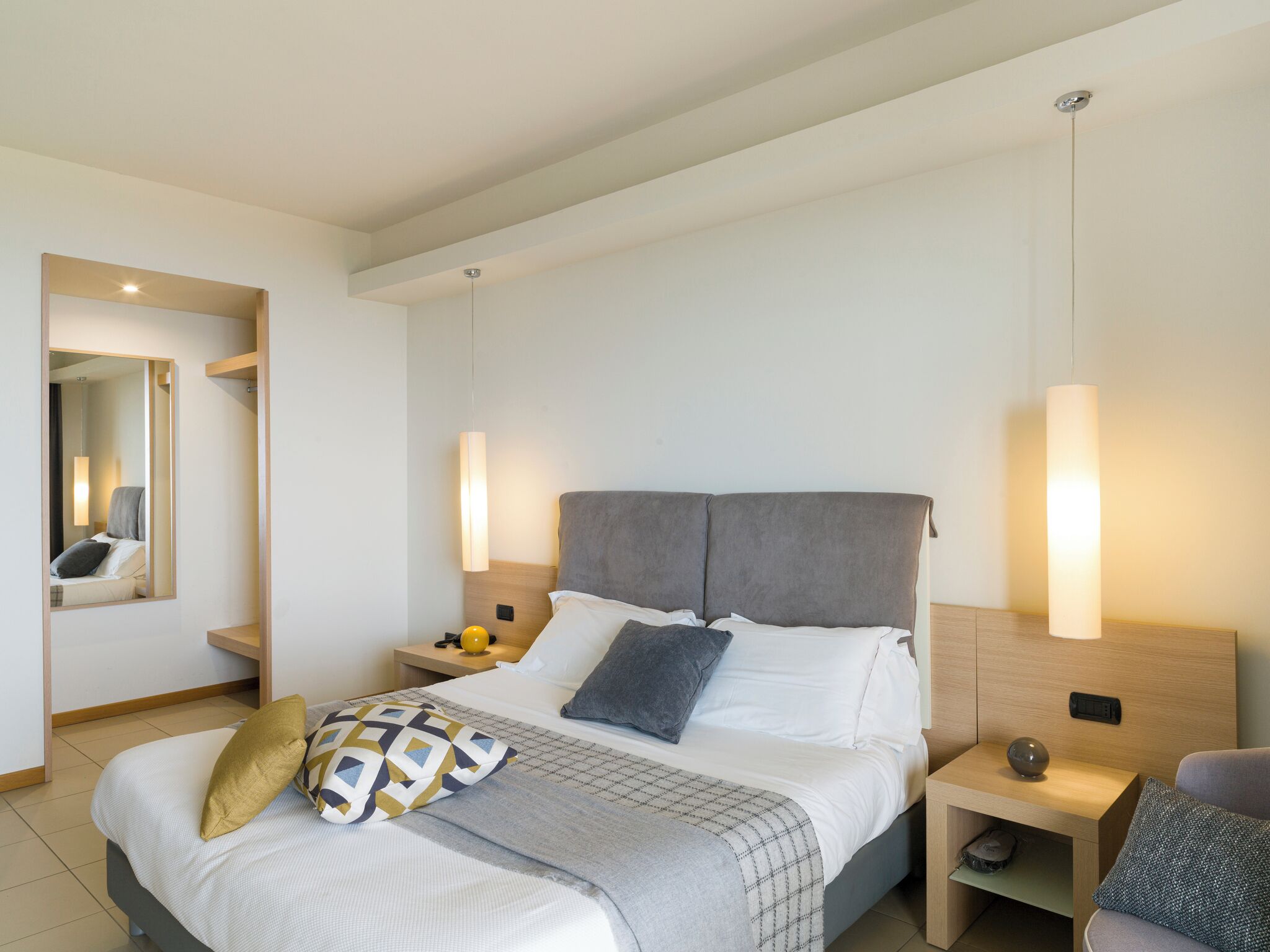 A modern hotel room with a large bed, bedside tables, a mirror, and wall-mounted lighting.