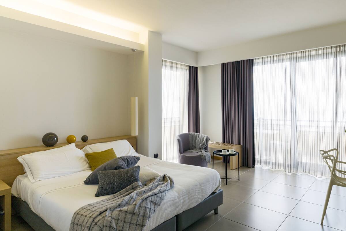 Una habitación de hotel moderna con una cama grande, una mesita auxiliar pequeña, una silla y vistas al exterior a través de cortinas traslúcidas.