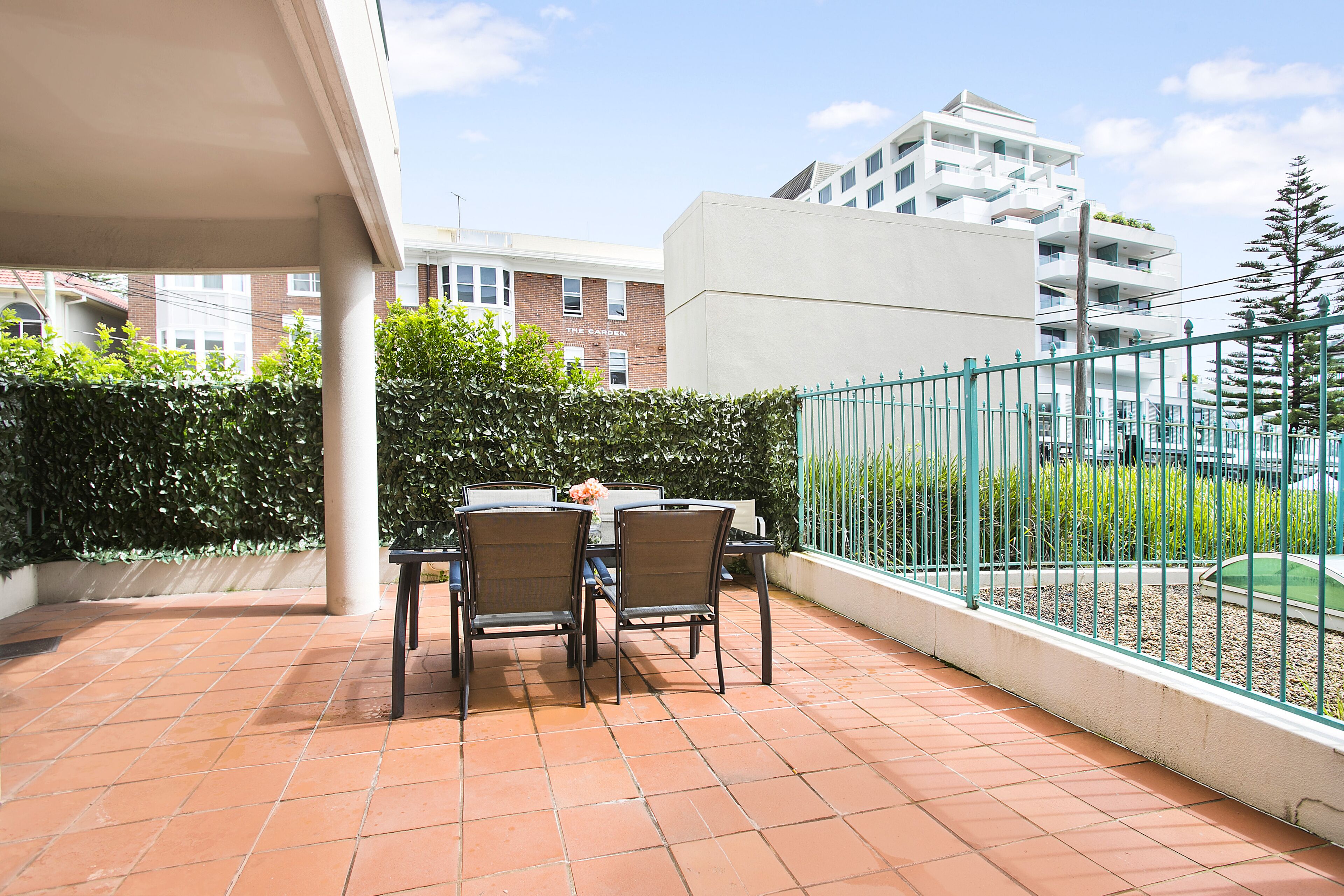 Una terraza con una mesa y sillas, piso de baldosas, cerca verde y un edificio al fondo.