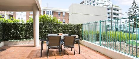 Una terraza con una mesa y sillas, piso de baldosas, cerca verde y un edificio al fondo.