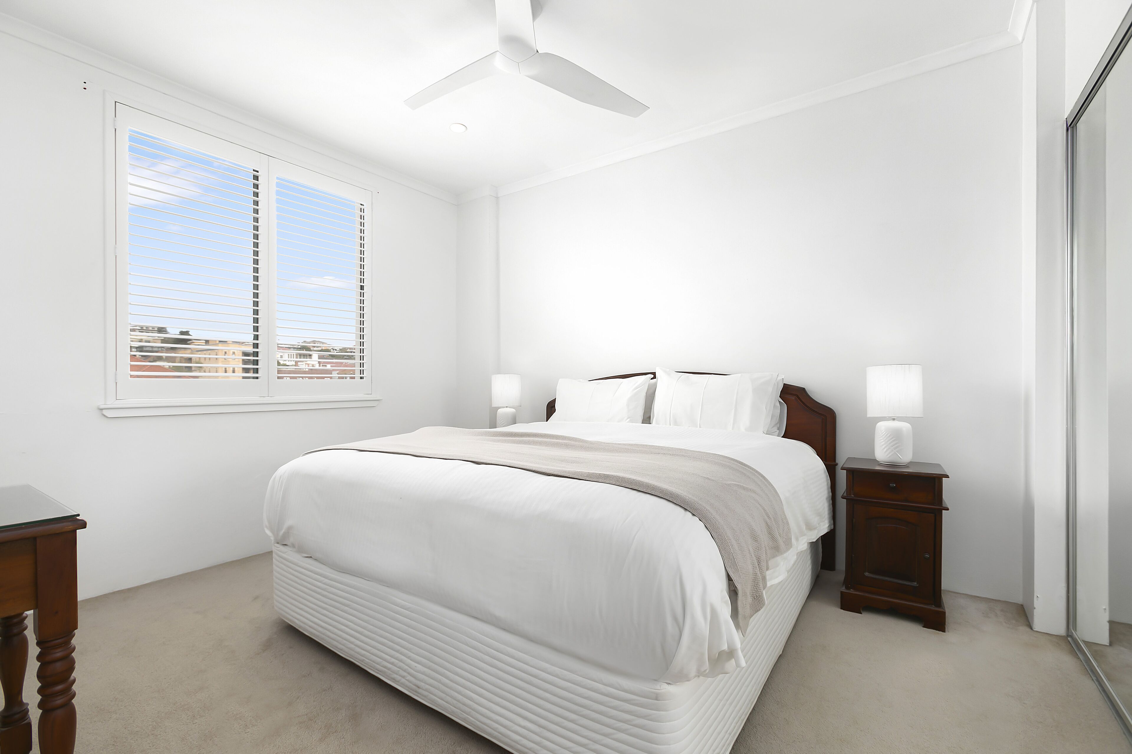 Un dormitorio con una cama grande, lámparas de noche, ventilador de techo y una ventana con persianas.